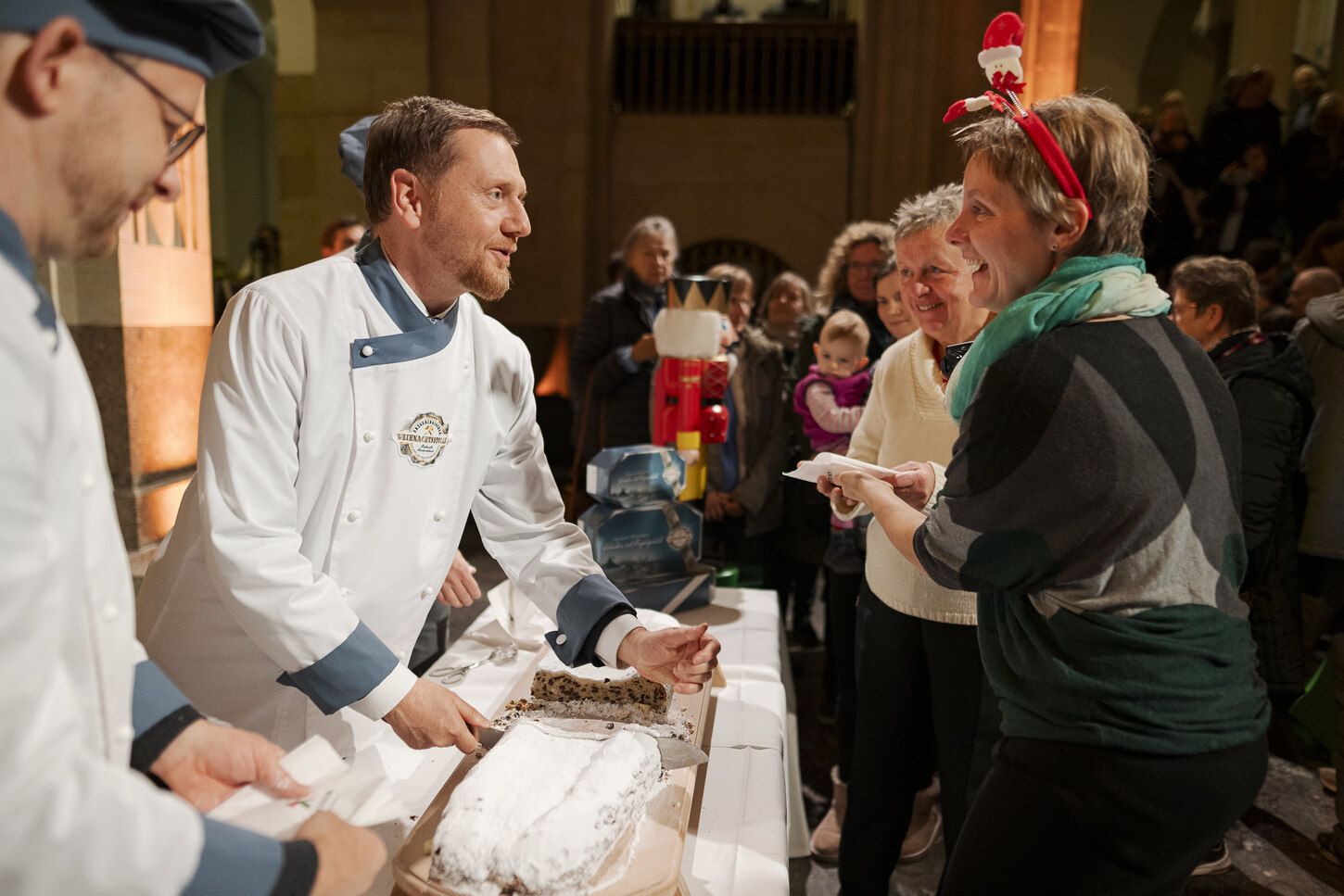 Ein Mann in Bäckeruniform teilt ein Stück von einem großen Stollen an eine Frau aus. Sie unterhalten sich.