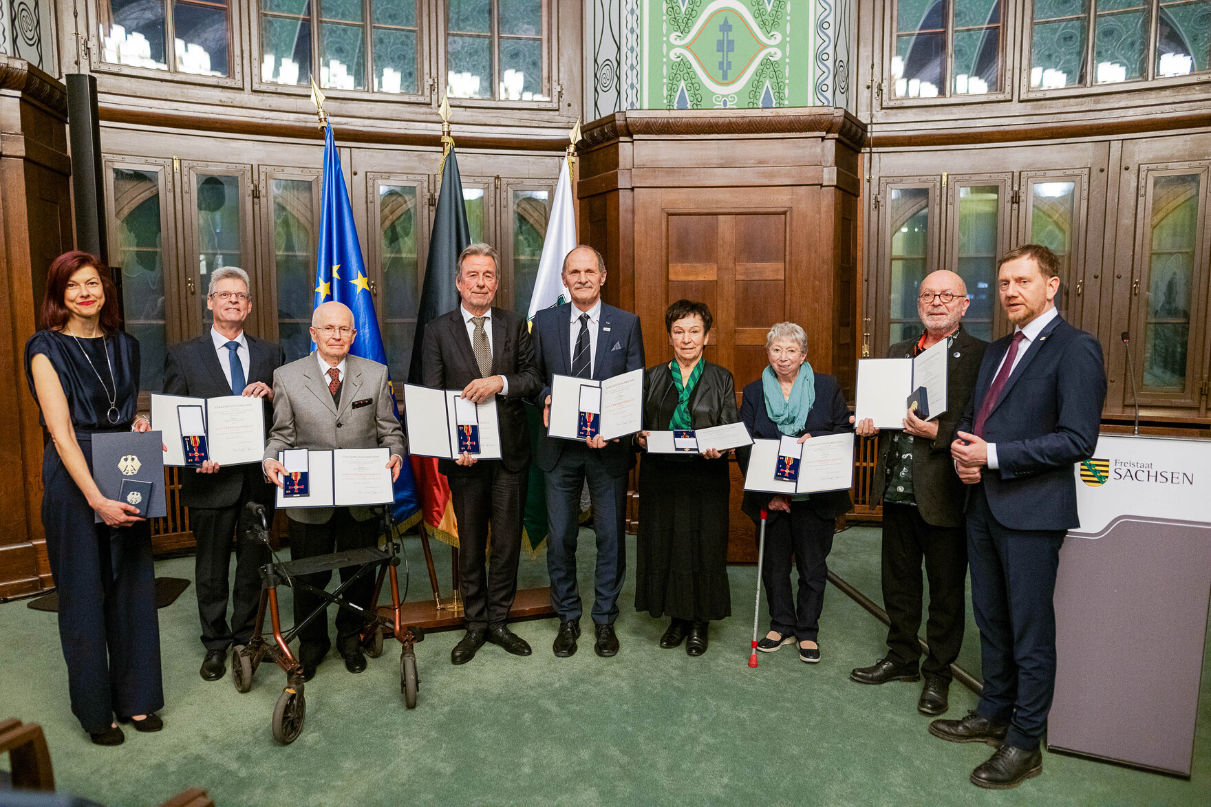 Männer und Frauen stehen in einem Saal zusammen und halten eine Urkunde in der Hand. Bildunterschrift: Verleihung des Bundesverdienstordens am 9. Dezember 2024 in der Sächsischen Staatskanzlei.