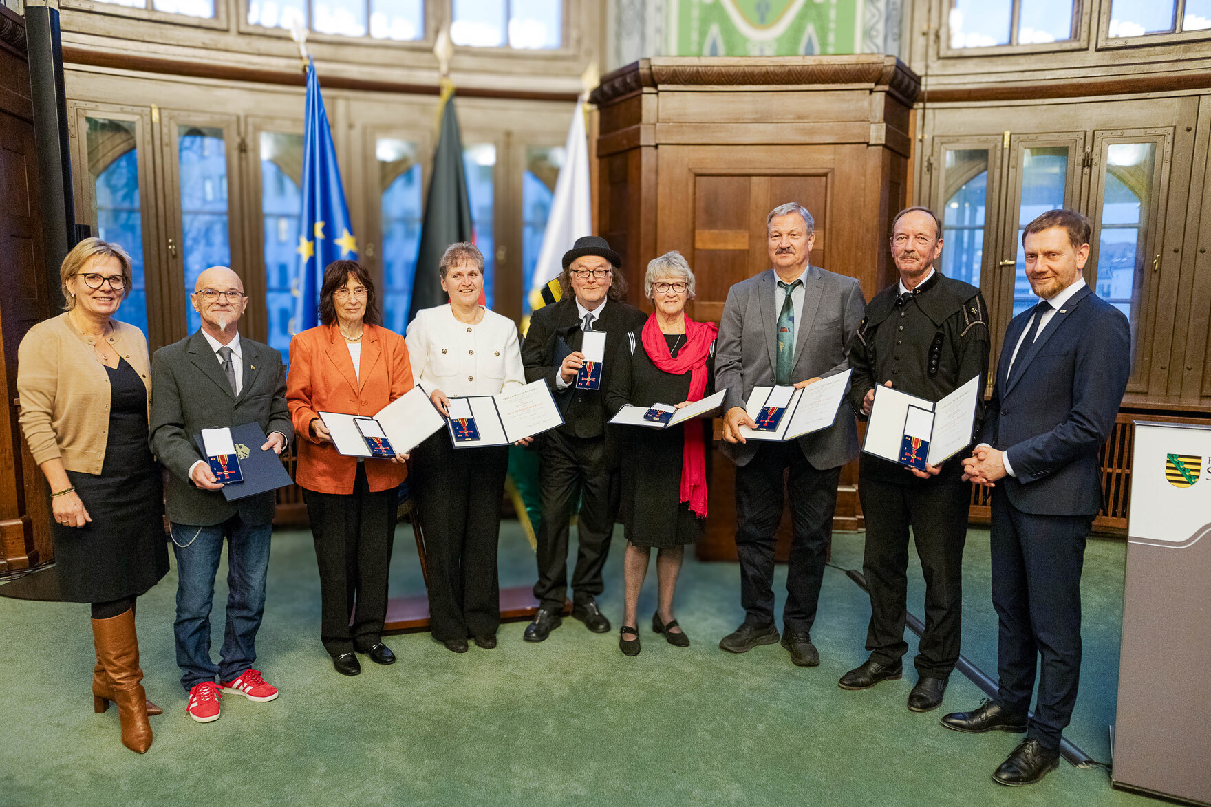 Männer und Frauen stehen in einem Saal zusammen und halten eine Urkunde in der Hand. Bildunterschrift: Verleihung des Bundesverdienstordens am 10. Dezember 2024 in der Sächsischen Staatskanzlei.