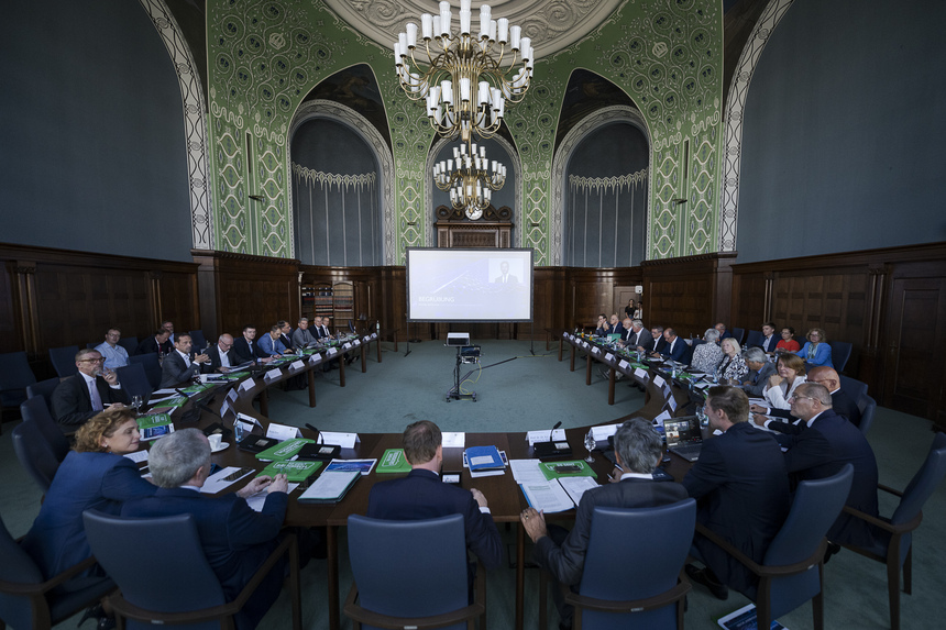 Männer und Frauen sitzen an einem großen Konferenztisch und blicken auf eine Leinwand.