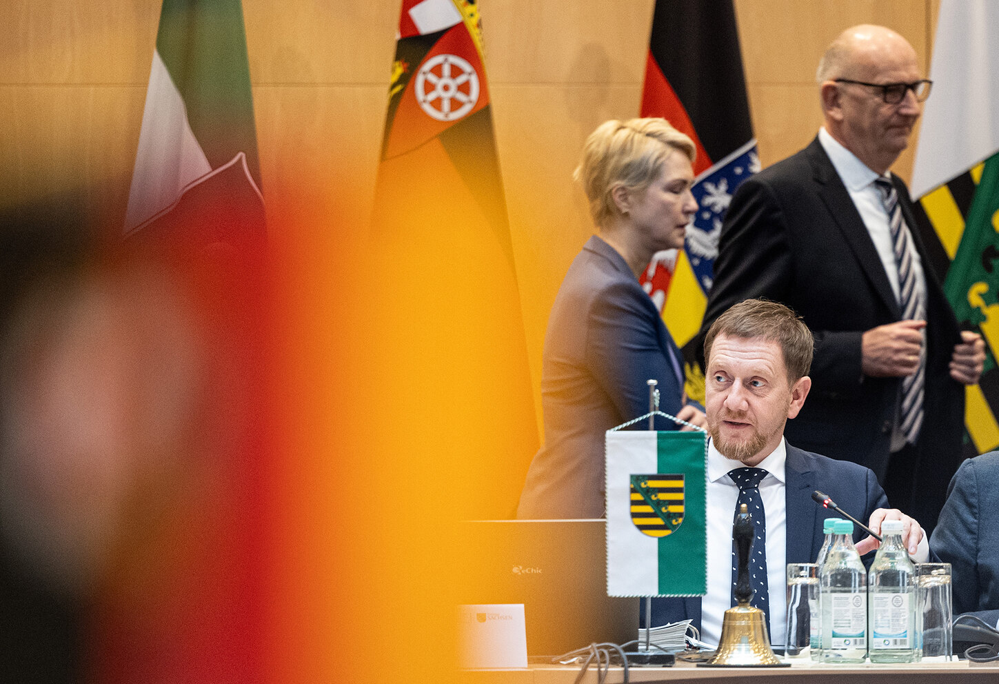 Ein Mann sitzt an einem Konferenztisch. Im Hintergrund sind weitere Personen und Flaggen zu erkennen.