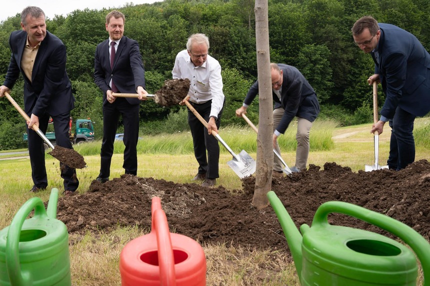 Männer schaufeln mit Spaten. Daneben stehen bunte Gießkannen.