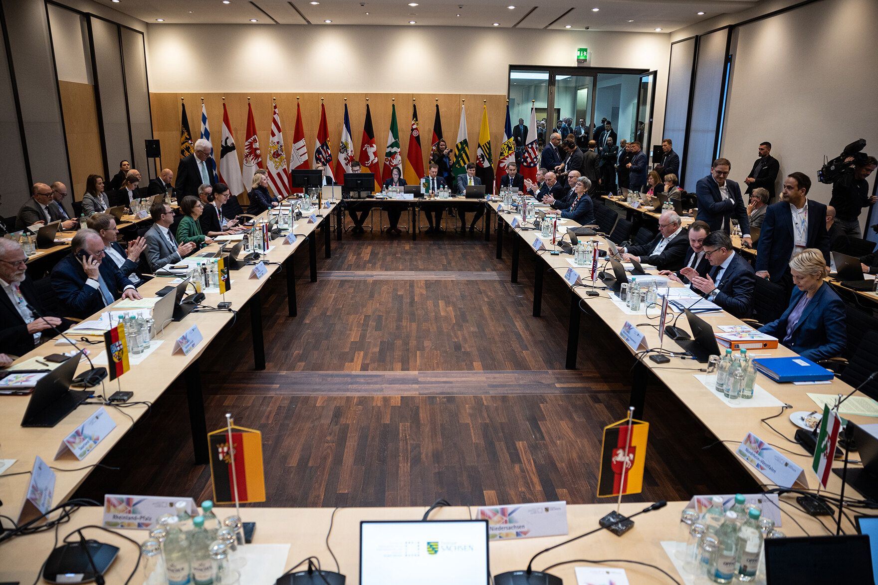 Männer und Frauen sitzen an einem Konferenztisch. Im Hintergrund stehen Fahnen der Bundesländer Deutschlands.